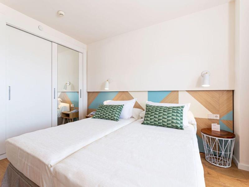 Nowoczesny pokój hotelowy z dwoma pojedynczymi łóżkami, geometrycznym zagłówkiem i małym stolikiem nocnym.