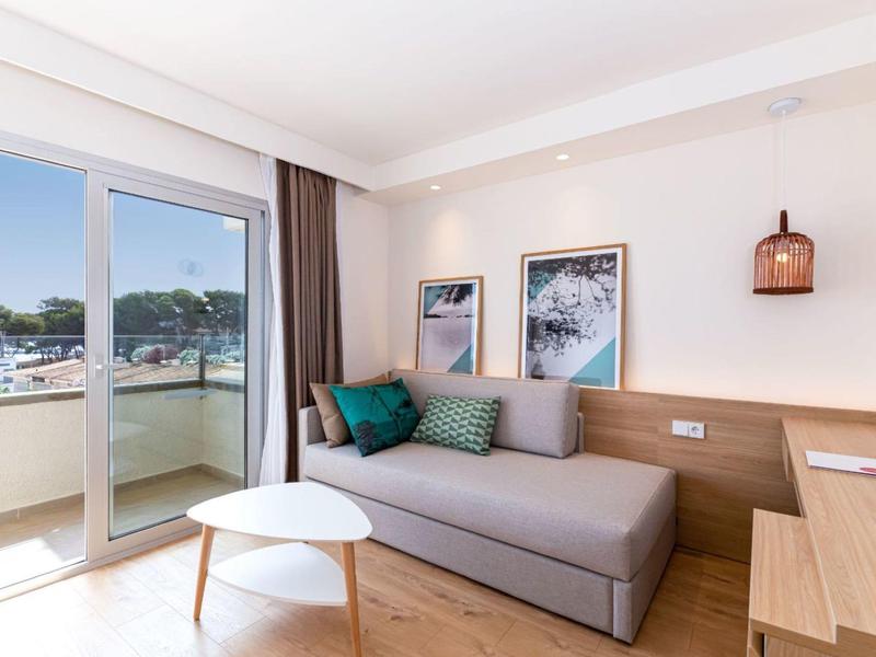 Jasno urządzony pokój hotelowy z sofą rozkładaną, stolikami i dostępem do balkonu.