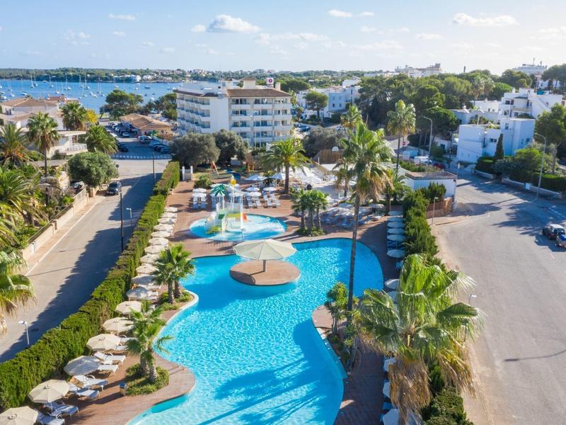 Widok na hotel z dużym basenem, parasolami i otaczającymi palmami.