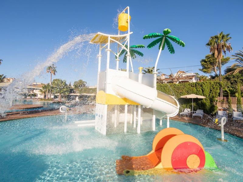 Park wodny z białą zjeżdżalnią, palmami i brodzikiem w słoneczny dzień.