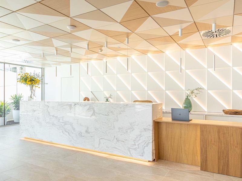 Jasne lobby hotelowe z marmurowym i drewnianym biurkiem oraz geometrycznym sufitem.