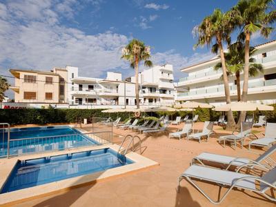 Terrasse mit Liegestühlen, Pool und Palmen vor weißem, mehrstöckigem Hotelgebäude bei blauem Himmel.