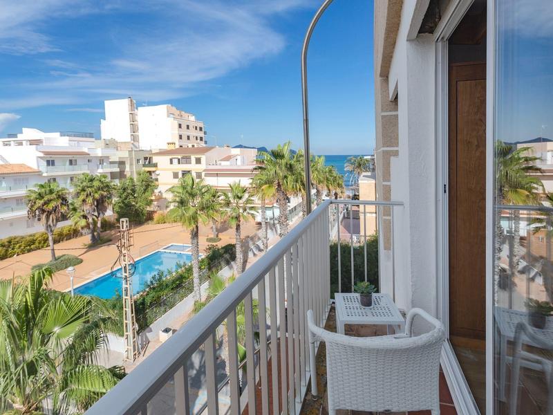 Balcón con sillas blancas, vista a jardín de palmeras, piscina y edificios blancos bajo cielo azul.
