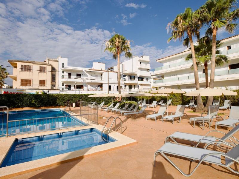 Área de piscina del hotel con tumbonas, palmeras y edificios blancos de varios pisos bajo cielo azul.