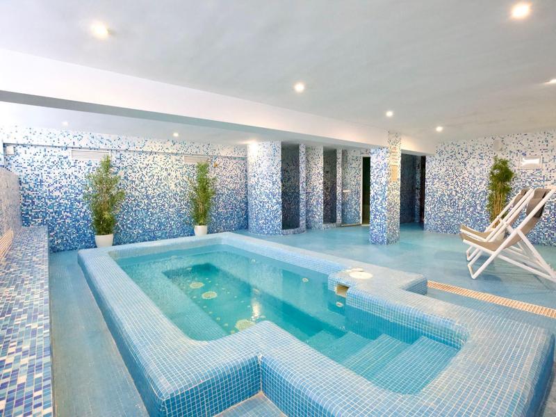 Piscina interior luminosa con azulejos mosaicos azules y tumbonas en un área de bienestar.