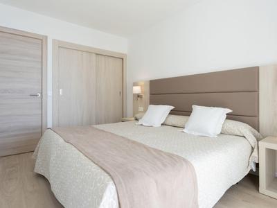 Světý hotelový pokoj s manželskou postelí, dvěma bílými polštáři, dřevěným obložením a nočním stolkem s lampou.