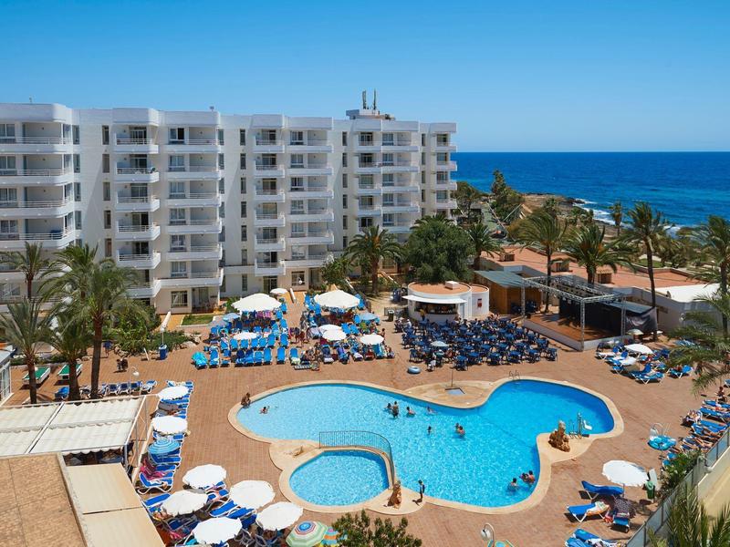 Hotel s bílými budovami, bazénem, slunečníky a výhledem na moře pod jasnou oblohou.