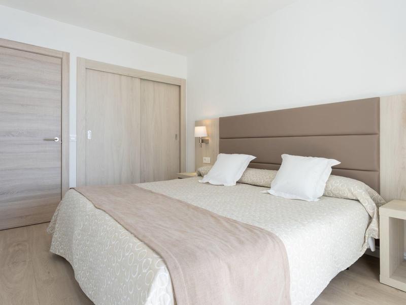 Světý hotelový pokoj s manželskou postelí, dvěma bílými polštáři, dřevěným obložením a nočním stolkem s lampou.