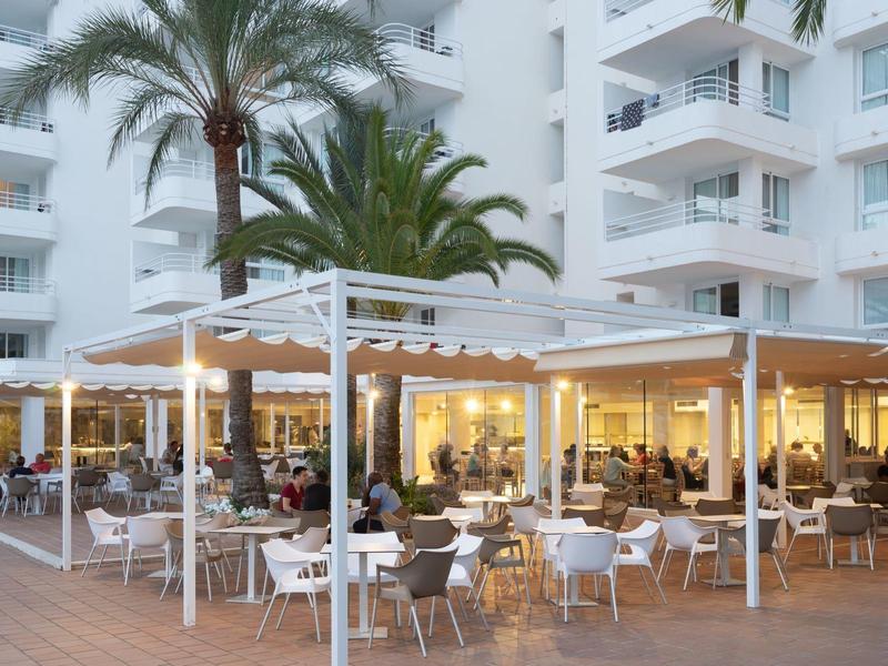 Venkovní restaurace s pergolou a židlemi před vícepatrovou hotelovou budovou.