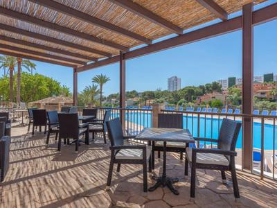 Zastřešená venkovní terasa se stoly a židlemi vedle bazénu s výhledem na palmy a budovy.