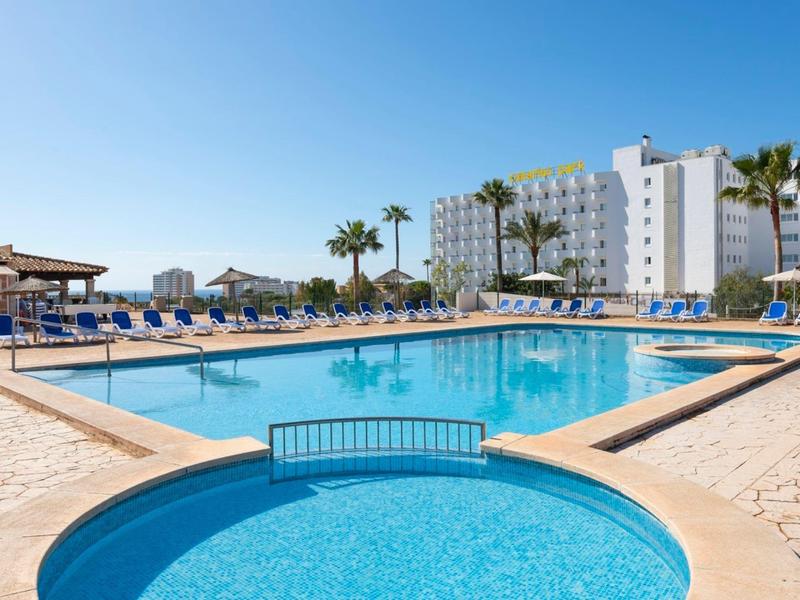 Grande piscina d'hotel con lettini blu e palme sotto un cielo limpido.