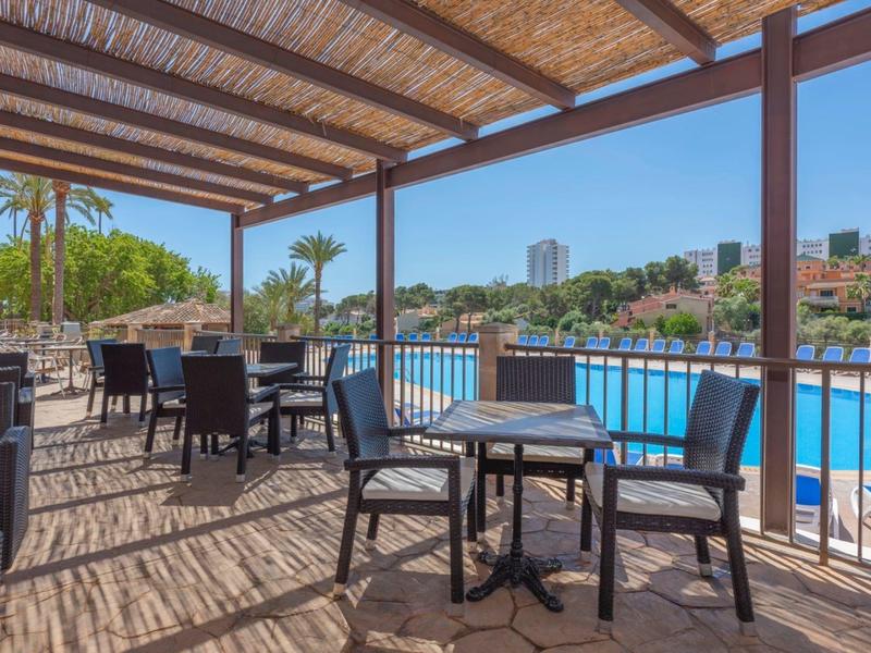 Terrazza ombreggiata con tavoli e sedie affacciata su una piscina con palme.