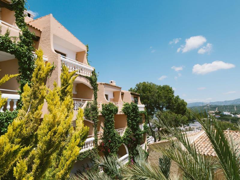 Mehrstöckige Wohnhäuser mit Balkonen, umgeben von grünen Bäumen und vor blauem Himmel mit Wolken.