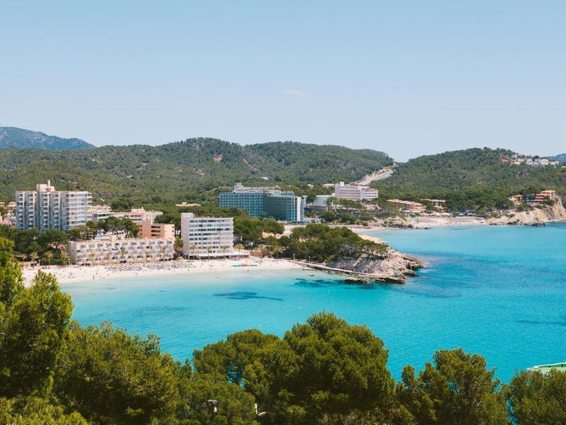 Küste mit türkisfarbenem Meer, weißem Sandstrand, Hotels und grünen Bergen im Hintergrund.
