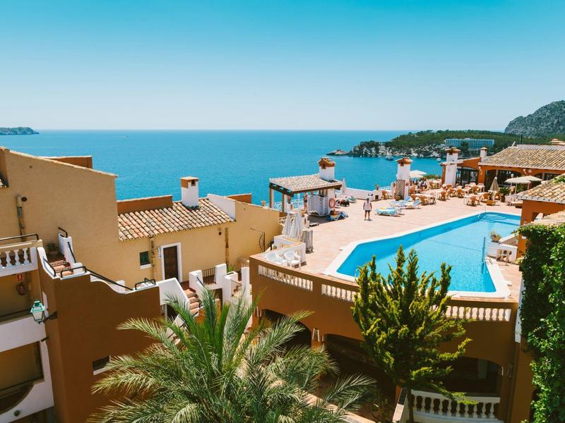 Blick auf ein Hotel mit Pool, umgeben von Palmen, mit Meer und Insel im Hintergrund bei klarem Himmel.