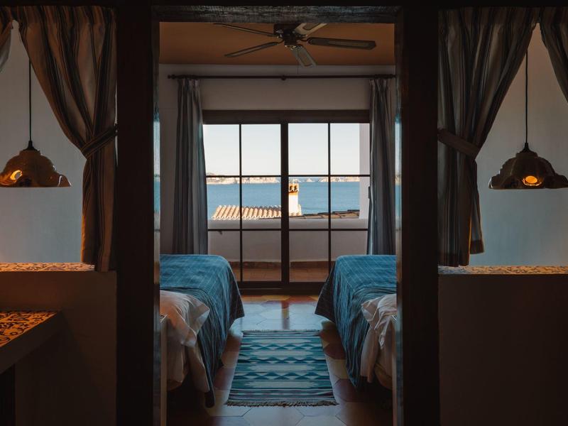 Schlafzimmer mit zwei Betten, Teppich, hängenden Lampen und Balkon mit Meerblick.