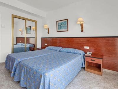 Pokój hotelowy z dwoma łóżkami, stolikiem nocnym i lampami ściennymi w jasnym pomieszczeniu.