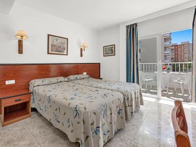 Pokój hotelowy z dwoma pojedynczymi łóżkami, stolikiem nocnym, lampami ściennymi i balkonem z widokiem na miasto.