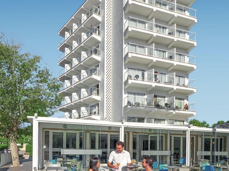 Moderner, mehrstöckiger Betonbau mit hellen Balkonen und Außenbereich mit Tischen und Stühlen.