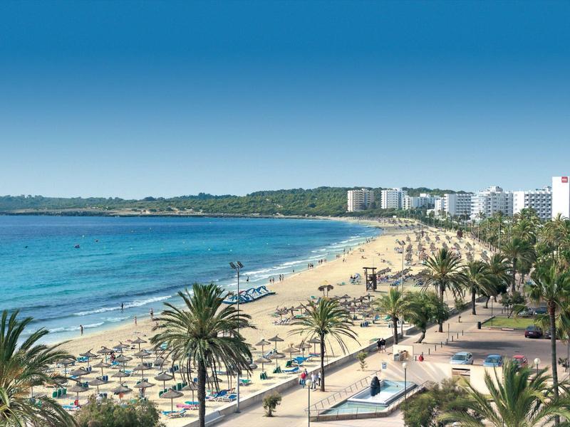 Strand mit Sand, Palmen und blauem Meer, im Hintergrund moderne weiße Gebäude und blauer Himmel.