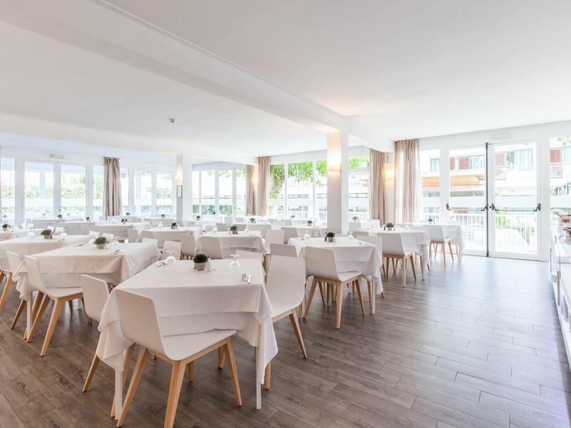 Großer, heller Speisesaal mit weißen Tischdecken und Holzstühlen, vielen Fenstern und dunklem Boden.