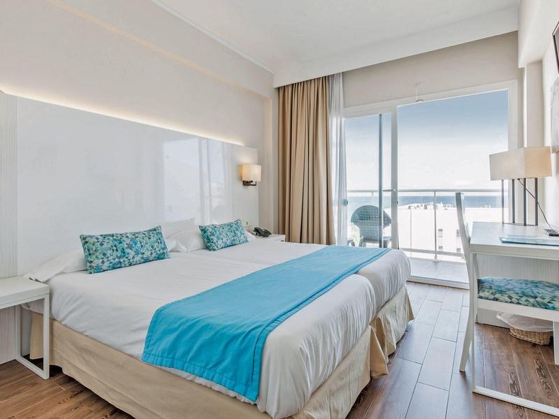 Helles Schlafzimmer mit Doppelbett, blauen Akzenten und Balkon mit Meerblick.