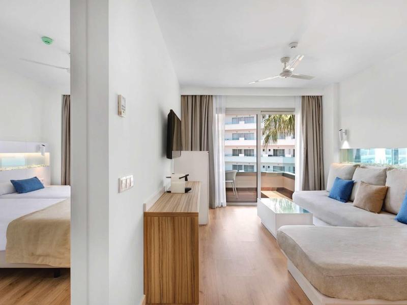 Jasne pokoje hotelowe z drewnianą podłogą, białymi ścianami, sofami i dostępem do balkonu.