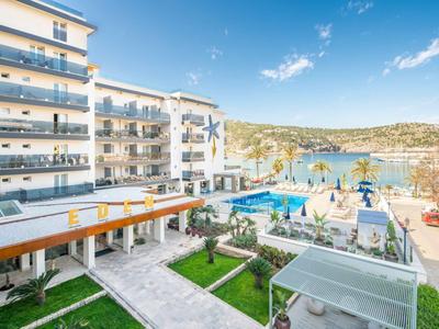 Moderner Hotelkomplex mit Pool, gepflegten Grünflächen und Blick auf das Meer und eine Küstenstadt.