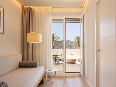 Helles Hotelzimmer mit Sofa, Stehlampe und Balkon mit Aussicht auf Palmen.