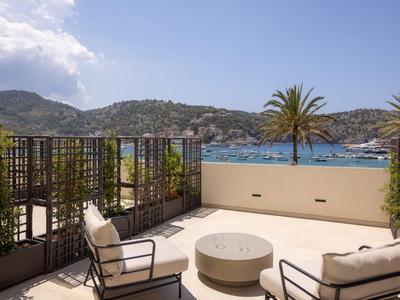 Terrasse mit Sitzmöbeln, Blick auf das Meer, Palmen und Hügel bei klarem Himmel.