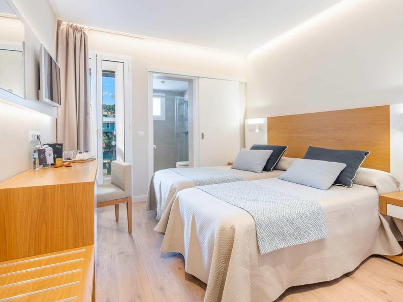 Helles Doppelzimmer mit zwei Betten, Holzverkleidung, Schreibtisch und Balkon mit Blick nach draußen.