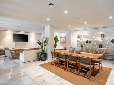 Moderne hotel lobby met een lange houten tafel, stoelen en een bankhoek met tv.