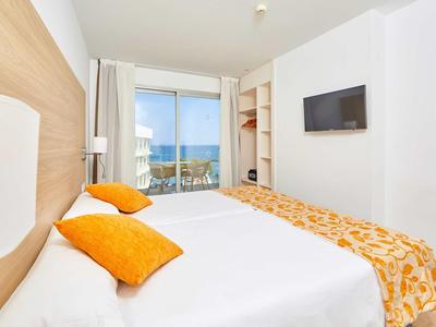 Helles Schlafzimmer mit Doppelbett, weißen Wänden, orangenen Kissen und Meerblick durch Fenster.