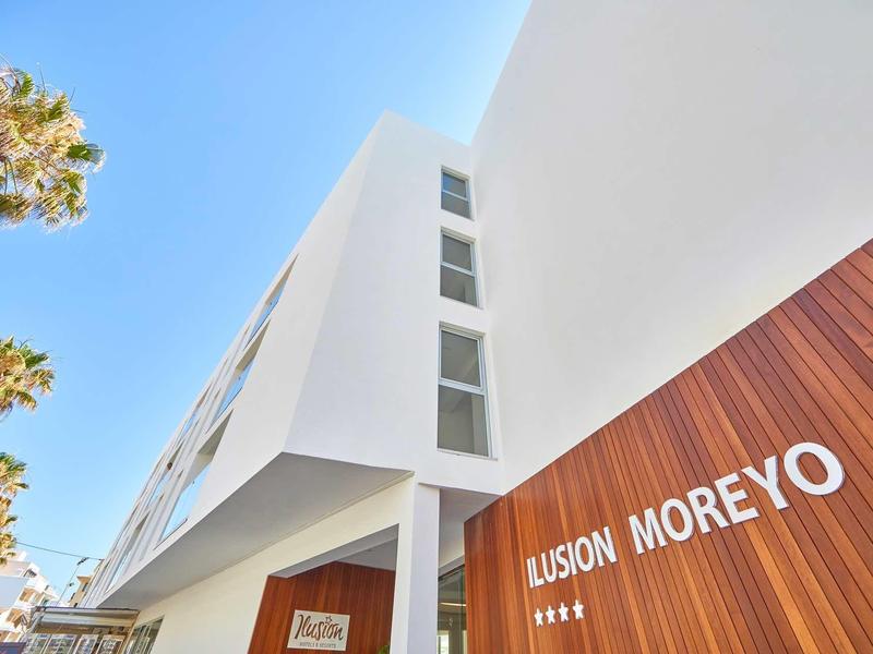 Moderne weiße Fassade mit Holzverkleidung und Schriftzug "ILUSION MOREYO" unter blauem Himmel.