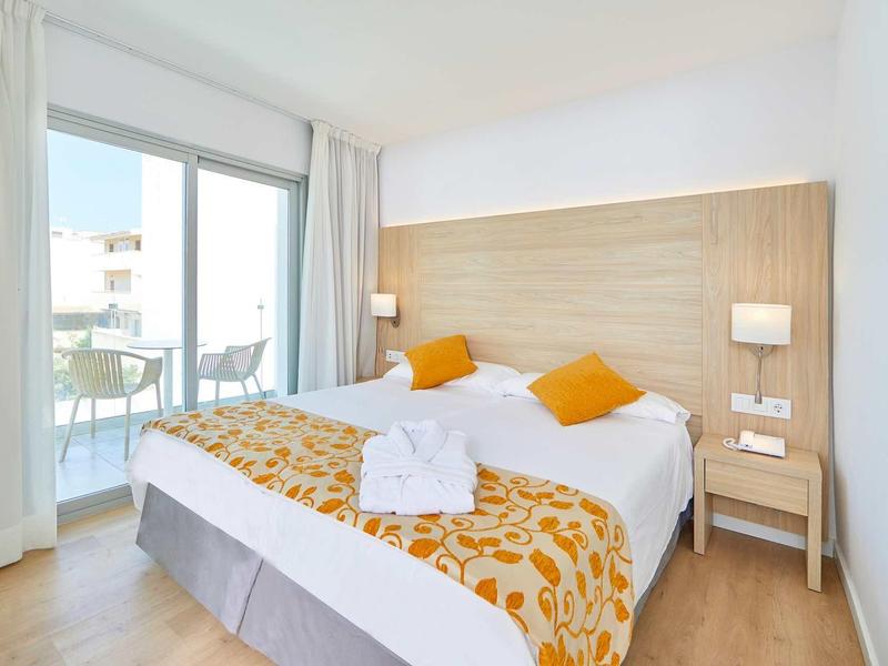 Helles Schlafzimmer mit Doppelbett, Holzkopfteil, orangen Kissen, Decke und Balkon mit Sitzgelegenheit.