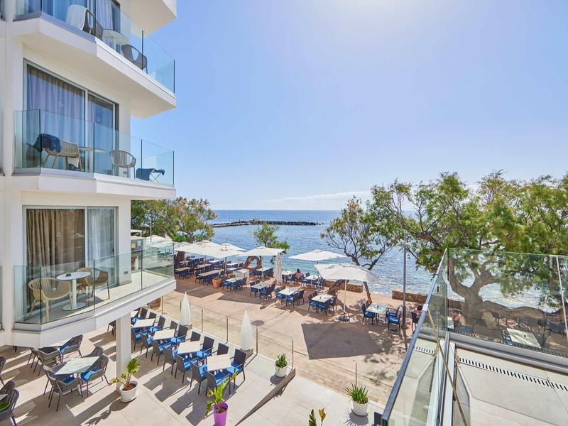 Moderner Hotelbereich mit Terrasse, Sonnenliegen, Bäumen und Blick aufs Meer bei klarem Himmel.