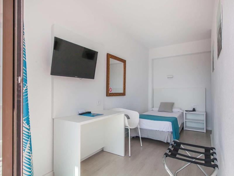 Mały jasny pokój z pojedynczym łóżkiem, biurkiem, krzesłem i telewizorem na ścianie.