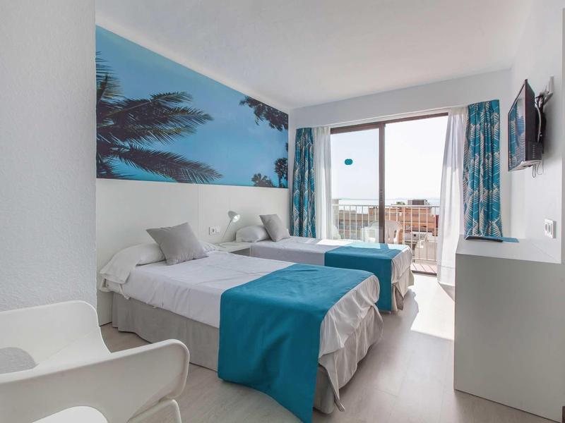 Jasny pokój hotelowy z dwoma łóżkami jednoosobowymi, niebieskimi kocami i balkonem z miejscami do siedzenia.