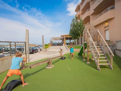 Kinder spielen auf dem künstlichen Rasen neben einem modernen Wohngebäude bei blauem Himmel.