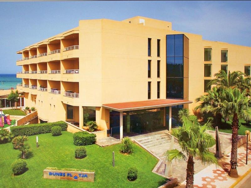 Gelbes, mehrstöckiges Gebäude neben Strand mit Palmen und gepflegtem Rasen unter blauem Himmel.