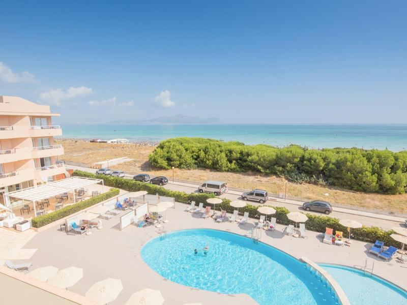 Hotel mit rundem Pool, Liegestühlen, Palmen und Blick auf den Strand und das blaue Meer.
