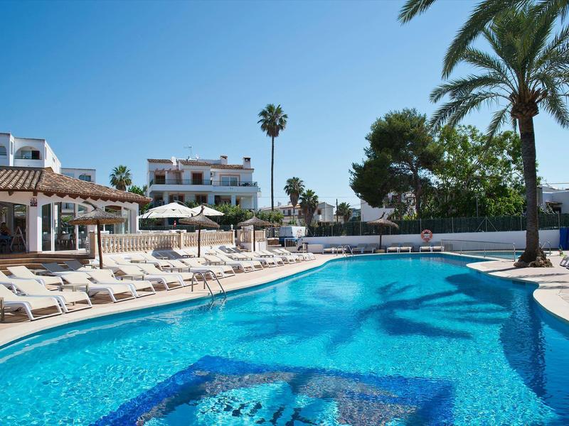 Outdoor pool area with sun loungers, umbrellas, and palm trees under a clear blue sky.