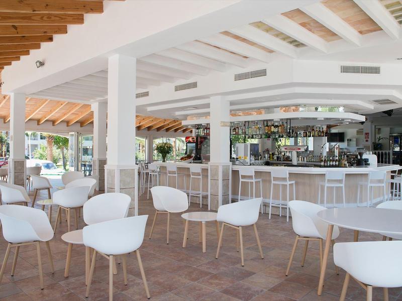 Spacious hotel bar area with white chairs, wooden tables, and a long counter under a white roof.