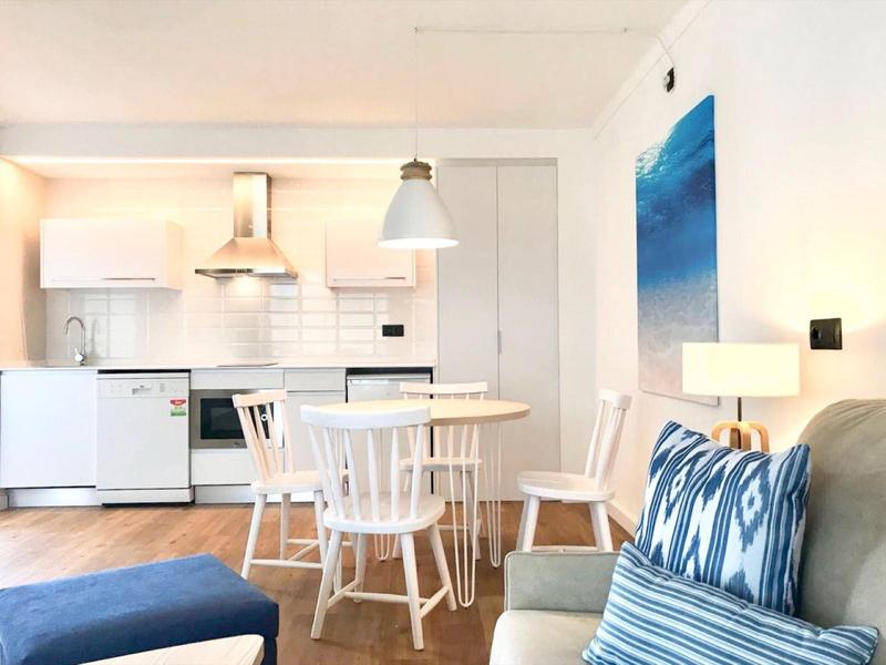 Cozy dining area with white wooden chairs and table, next to a modern kitchen and sofa with blue patterned pillows.