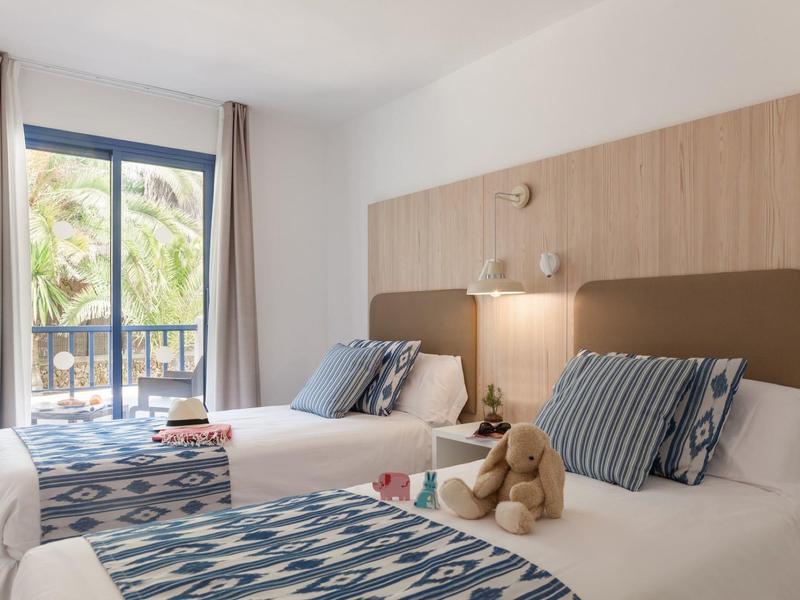 Hotel room with two single beds, blue patterned bed runners, striped pillows, and a teddy bear.