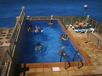 Rechteckiger Pool mit blauen Wasser, umgeben von beige Fliesen und mehreren Sonnenbadenden.