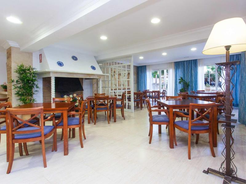 Heller Speiseraum mit Holztischen, blauen Stühlen, Kamin und Pflanzen, gut beleuchtet durch Deckenlichter.