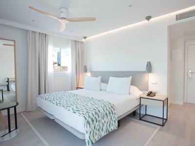 Helles, modernes Schlafzimmer mit großem Bett, weißen Wänden, Nachttisch, Lampe und Deckenventilator.