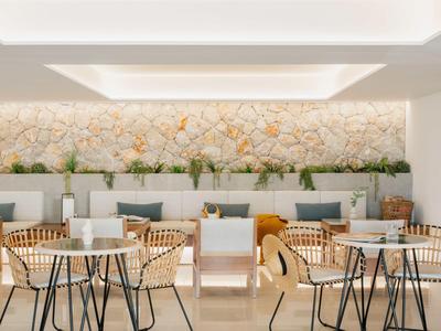 Moderne, lichte hotel lounge met rotan stoelen, witte tafels en groene planten langs de muur.