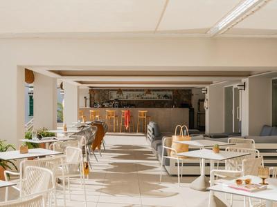 Lichte buitenruimte van een hotel met moderne tafels en stoelen, met een bar op de achtergrond.